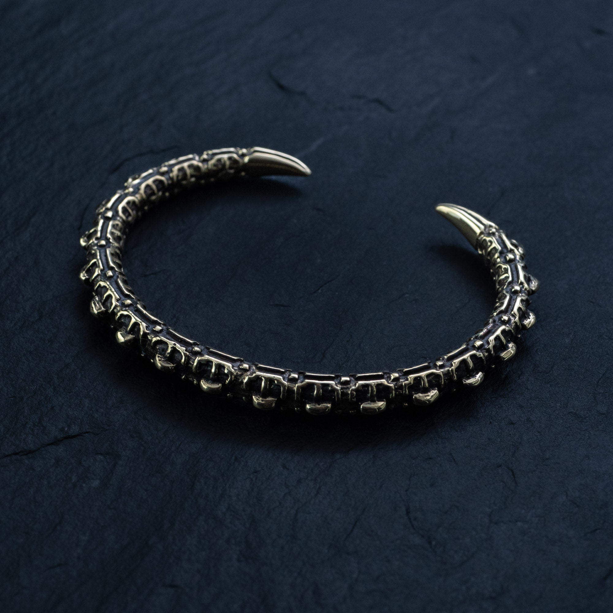 The SHIRO Men's Gold Cuff Bracelet by Kaya Movement, featuring an intricately designed reptile spine pattern and pointed ends resembling claws or fangs, rests on a dark, textured surface. This adjustable silver bracelet boasts a robust metallic structure and an ornate, almost gothic appearance.